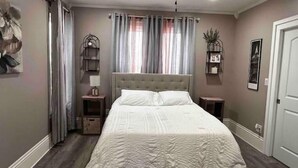 Un dormitorio con una cama grande, dos mesitas de noche, una mesita auxiliar, un ventanal con cortinas y estantes en la pared.