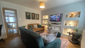 Una sala de estar con un sofá verde, un sofá marrón, un televisor en la pared y estanterías con libros y objetos decorativos.