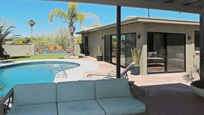 Un área de piscina moderna al aire libre con un espacio de estar cubierto, un patio con sillas reclinables y un edificio de cristal con un patio.