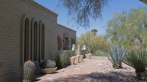 Un edificio de fachada de ladrillo, ventanas arqueadas y un camino pavimentado bordeado de cactus y plantas desérticas.