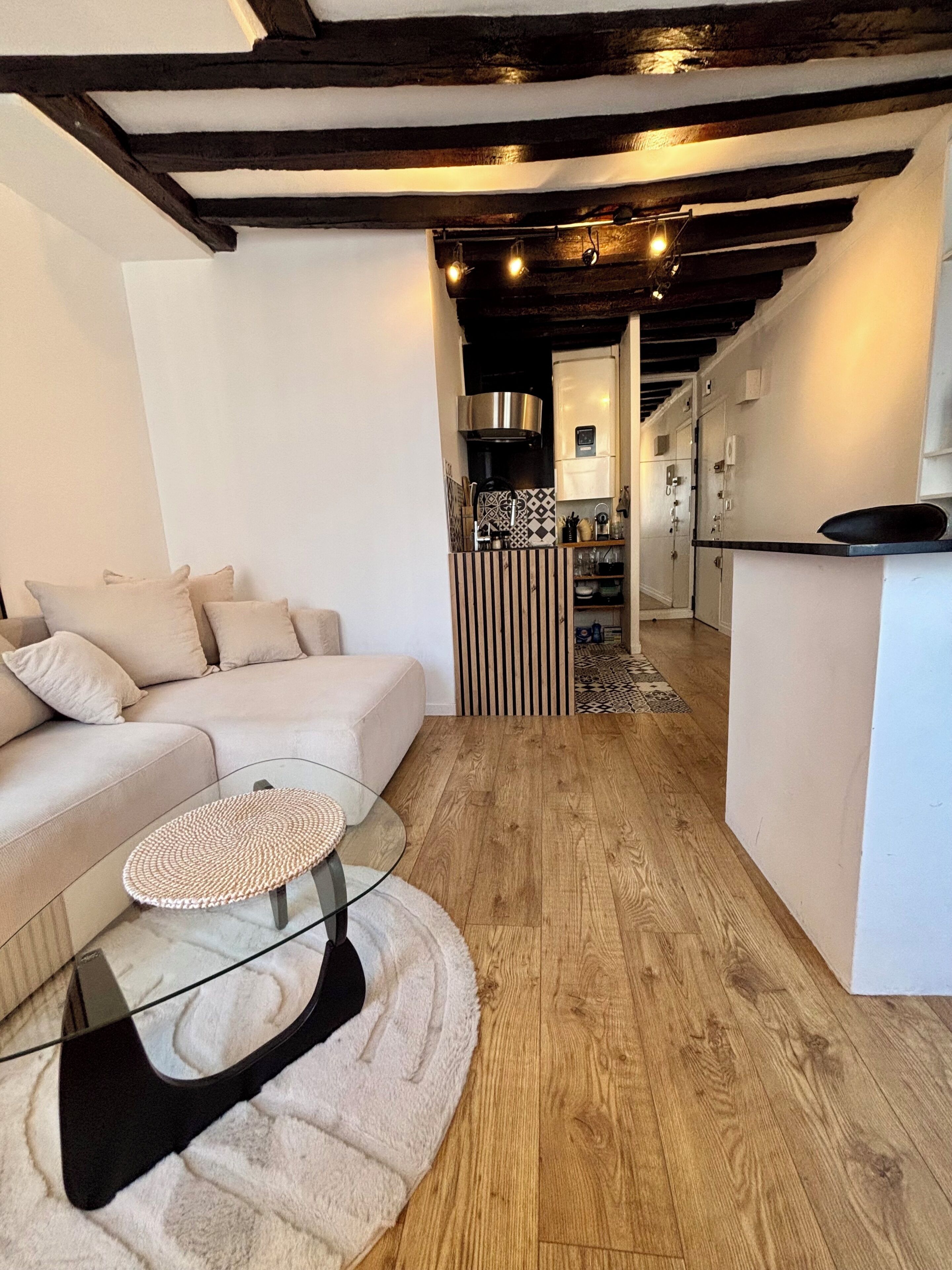 A modern living room with a sofa, a glass coffee table, and a kitchen area with a refrigerator and cabinets.
