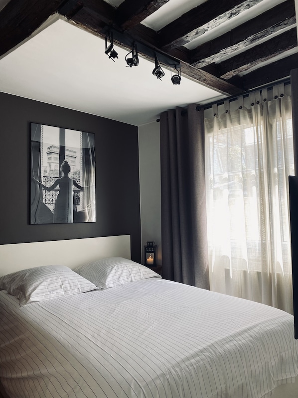 Une chambre à coucher avec un lit, un tableau encadré au mur, une table de chevet avec une lanterne et une fenêtre avec des rideaux.