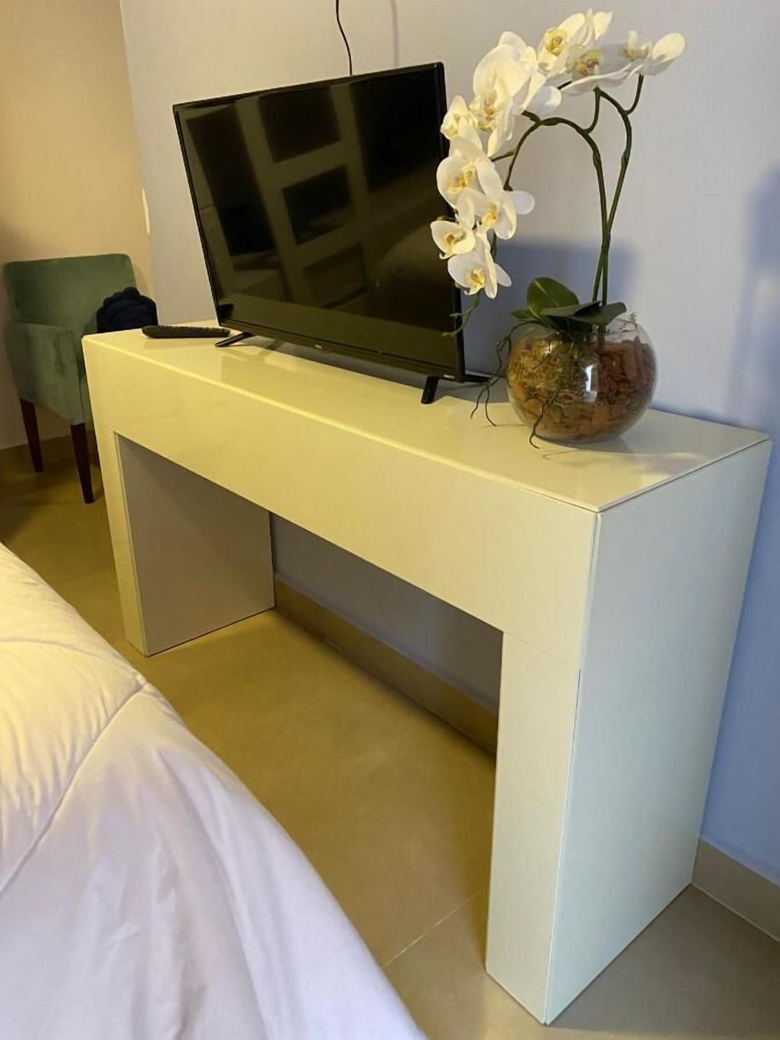 Une console blanche avec un téléviseur à écran plat, un vase contenant des orchidées blanches et une chaise verte, dans une chambre d’hôtel.