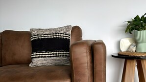 Un canapé en cuir marron agrémenté d’un coussin à motifs, une petite table d’appoint en bois sur laquelle est posée une plante en pot, et un tapis texturé sur un plancher en bois.