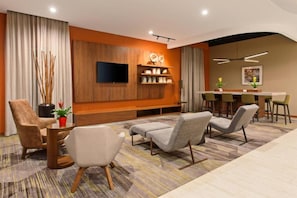 Una habitación de hotel moderna con una televisión de pantalla plana, una zona de estar con sofás y sillones, una mesa de comedor con sillas, y una pared decorativa con estanterías.