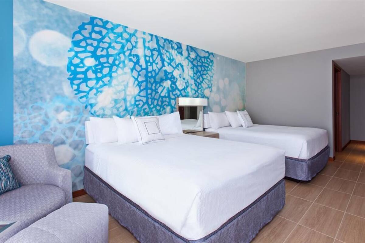 Habitación de hotel con dos camas, un sofá, una lámpara y una pared con un diseño abstracto azul.