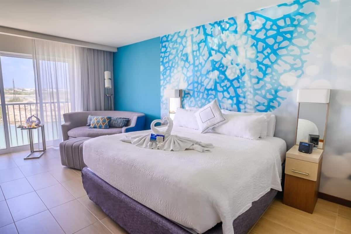 Habitación de hotel con una cama grande, un sofá, un balcón con vistas y un cuadro abstracto azul en la pared.
