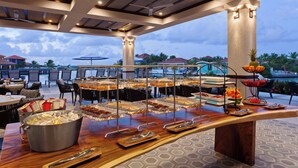 A buffet setup with various food items and utensils, a large pot of ice, and a view of the resort's pool and buildings.