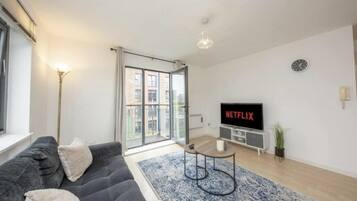 A modern living room with a grey sofa, a coffee table, a TV displaying 'NETFLIX', and a large window with a view of buildings.
