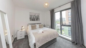 Un dormitorio moderno con una cama grande, mesitas de noche, un cuadro de una vista urbana y un amplio ventanal.