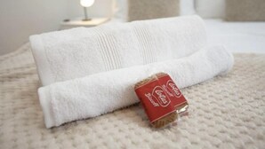 Una toalla blanca doblada con cuidado y un pequeño paquete rojo sobre una colcha beige con textura.