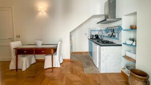 Una cocina compacta con estufa, lavamanos y salpicadero de azulejos blancos. Hay una mesa de comedor con sillas y una alfombra pequeña en el suelo.