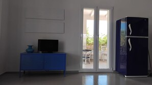 Una habitación con un armario azul, televisión, refrigerador y puerta corrediza de vidrio que da a un área exterior con una mesa y sillas.