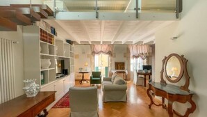 A spacious living room with a high ceiling, a staircase, and a large window. There are two sofas, a coffee table, and a wooden side table.