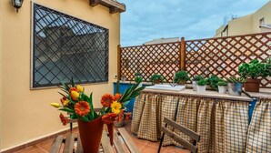 Eine Terrasse mit einer Holzbank, einem Tisch mit kariertem Tischläufer, Blumentöpfen und einem Fenster mit einem Metallgitter.