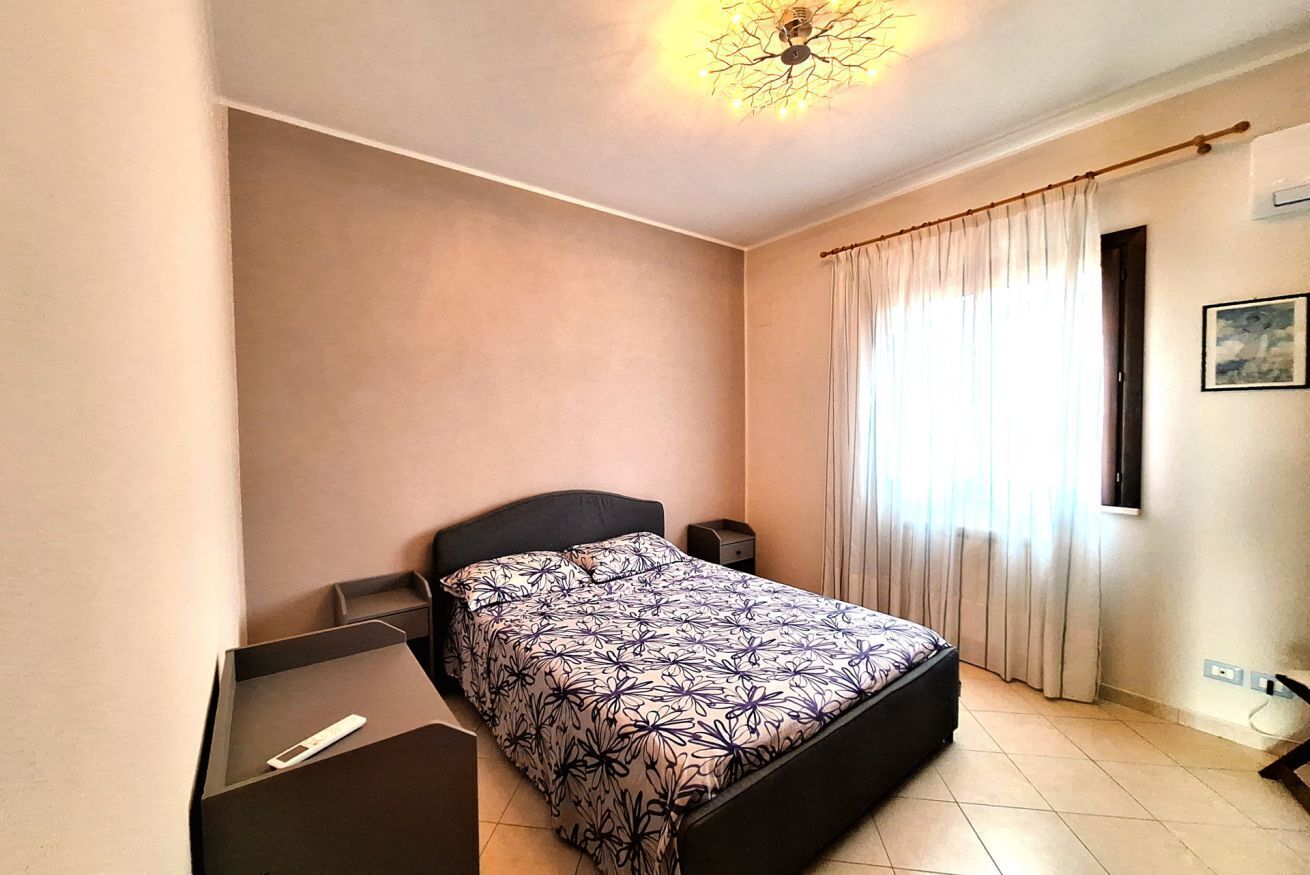 A bedroom with a bed, a bedside table, a nightstand, a window with curtains, and a wall-mounted air conditioning unit.