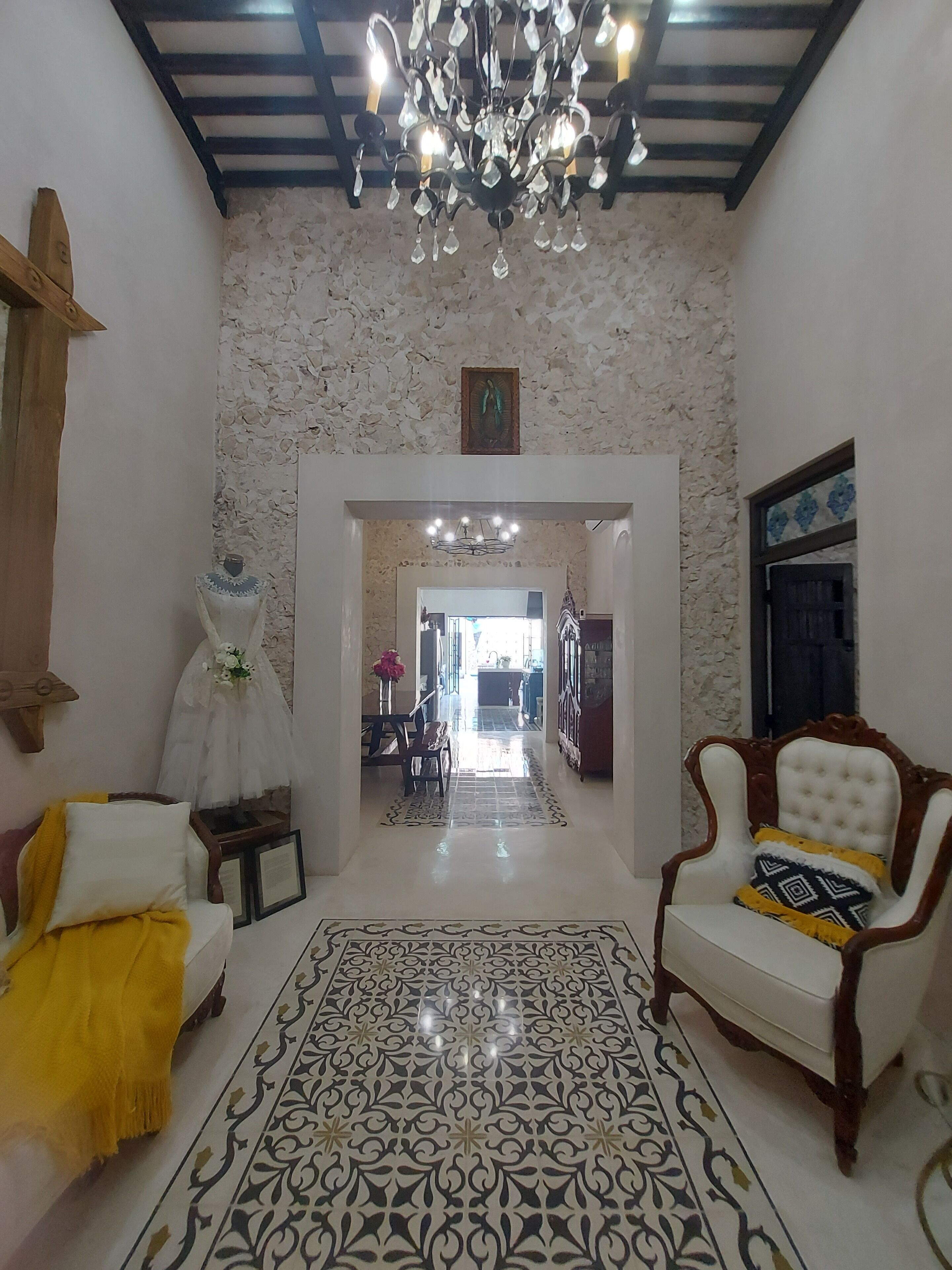 A spacious living room with a chandelier, a white armchair, a patterned rug, and a view into another room.