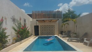 Piscina retangular com área coberta por sombra, rodeada por espreguiçadeiras e um muro decorativo com plantas.