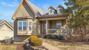 A two-story house with a front porch, a red door, and a small front yard with a stone planter and shrubs. - Dog-Friendly 2BR Lodge, Pool by Big Cedar (Hollister)
