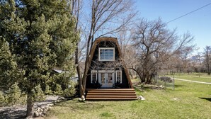 A small, A-frame house with a front porch, surrounded by trees and a grassy area. - The Cabin on Main St., A Quiet Getaway with whole family, History. Pet friendly. (Spring City)