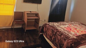 Un dormitorio con cama, escritorio, silla, televisor en la pared y ventana con cortinas.
