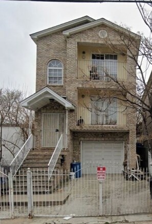 A two-story brick building with a garage door, a small front porch, and a fenced driveway.