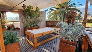 Eine Holzterrasse mit einer Bank und Pflanzgefäßen, mit Blick auf ein Schwimmbecken und Palmen.