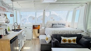 Una habitación compacta y bien iluminada con kitchenette, cama con ropa de cama estampada, sofá con almohadas de girasol y ventanas grandes con paraguas decorativos.