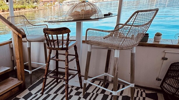 Una mesa con cubierta de vidrio y dos sillas de mimbre, con vista a un puerto deportivo con barcos y edificios al fondo.