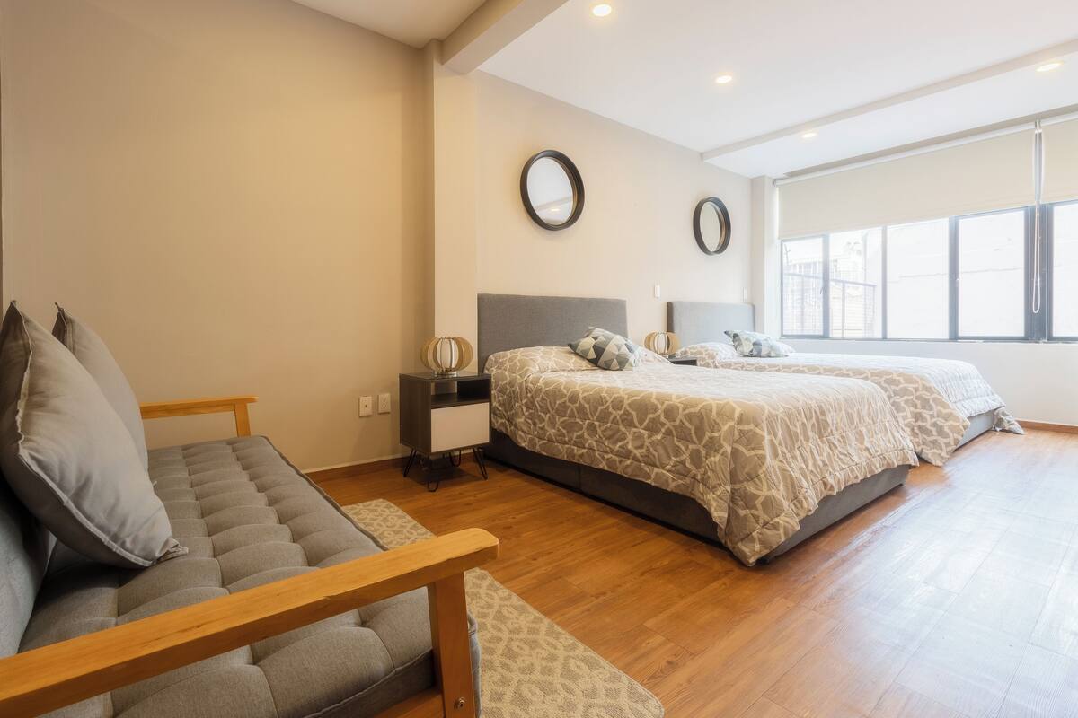 A bedroom with a sofa, two beds, a nightstand, and a large window.