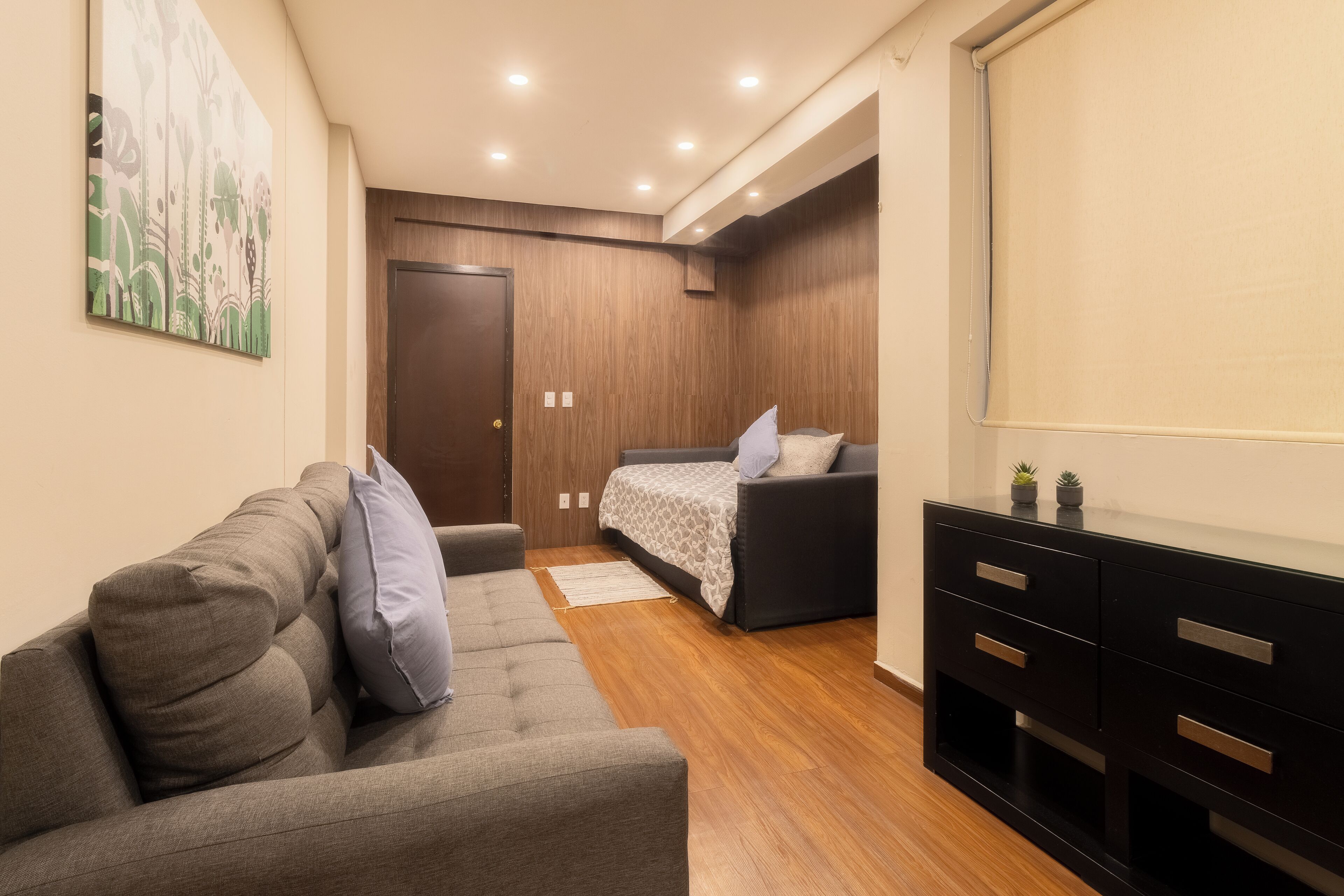A modern bedroom with a grey sofa, a bed with patterned bedding, a wooden wardrobe, and a wall-mounted artwork.