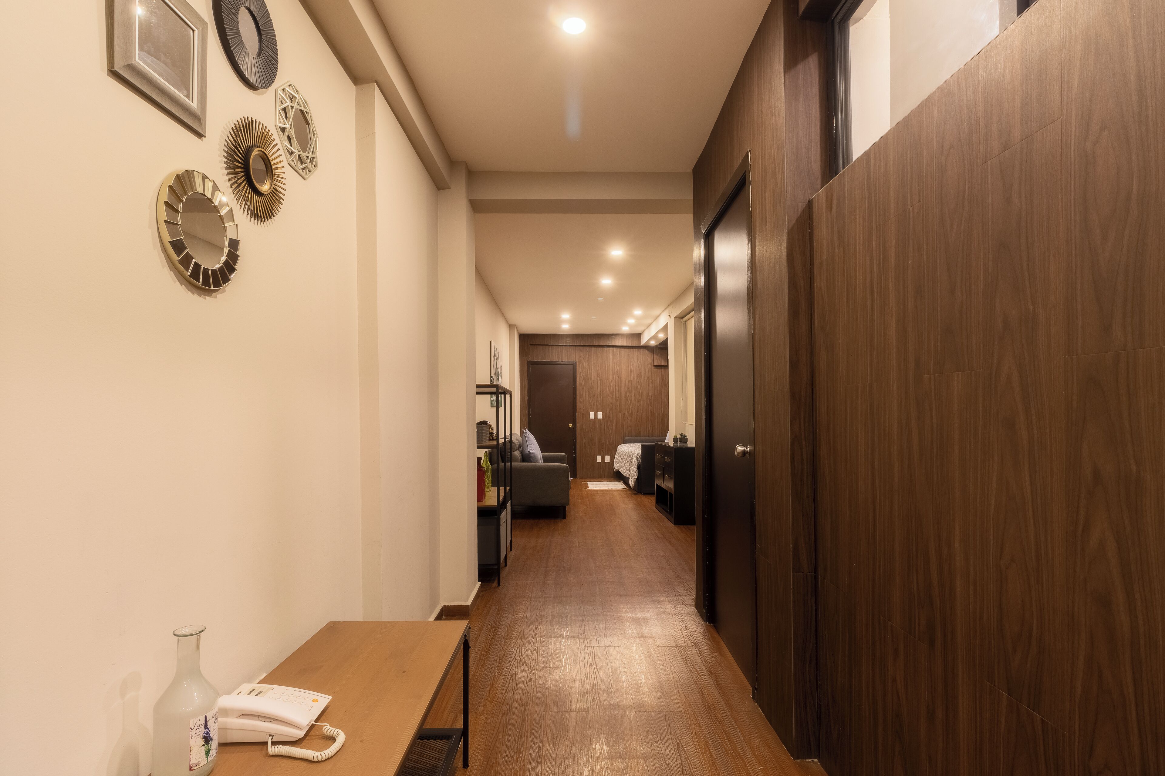 A hallway with a wooden floor, a table with a vase and a phone, and a door at the end.