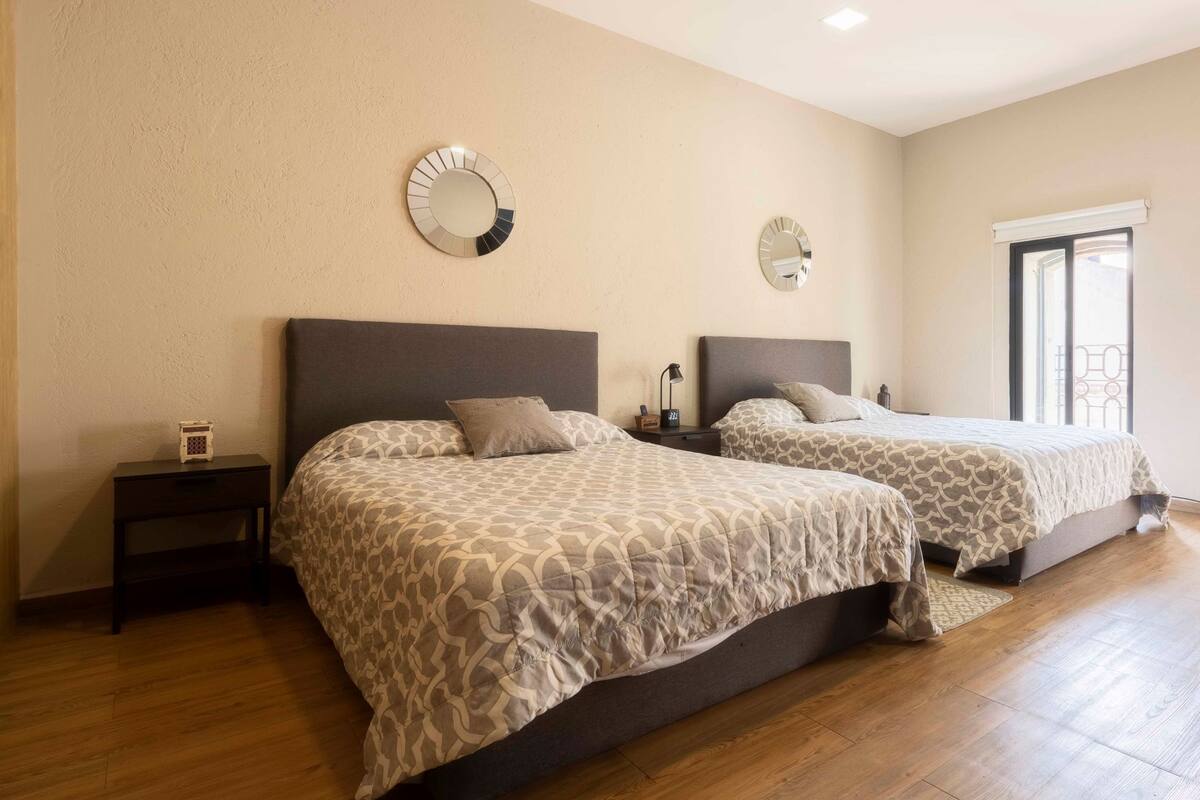 A hotel room with two beds, a small bedside table with a lamp, and a large window.
