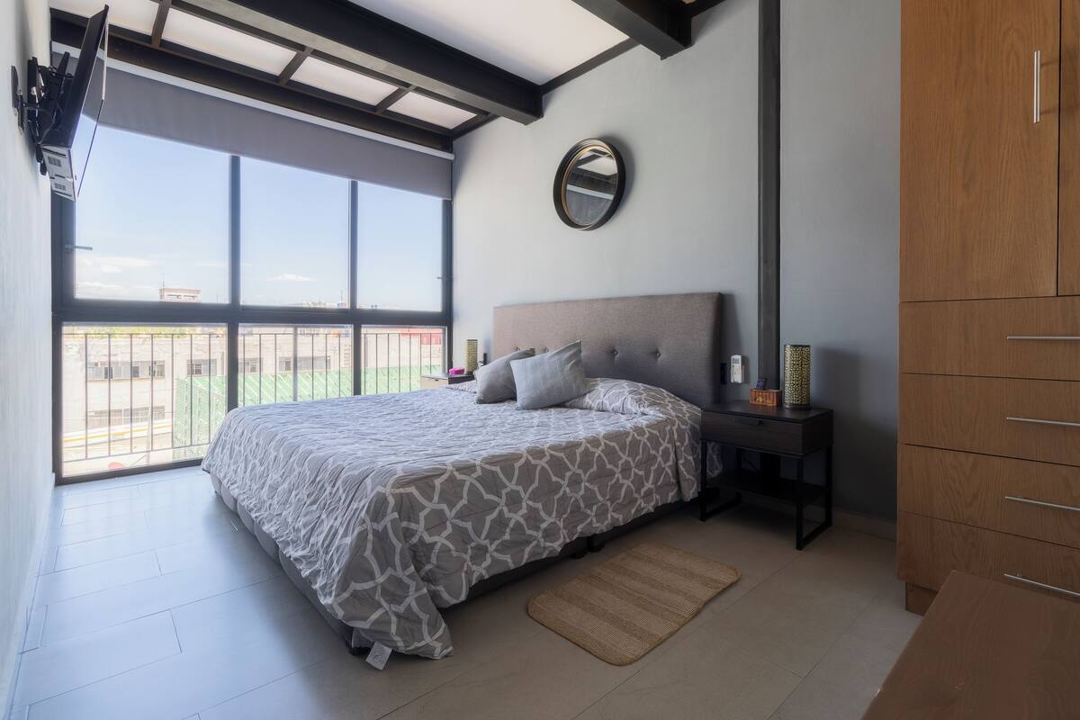 A bedroom with a large bed, a nightstand, a wardrobe, a mirror, and a view of the outside.