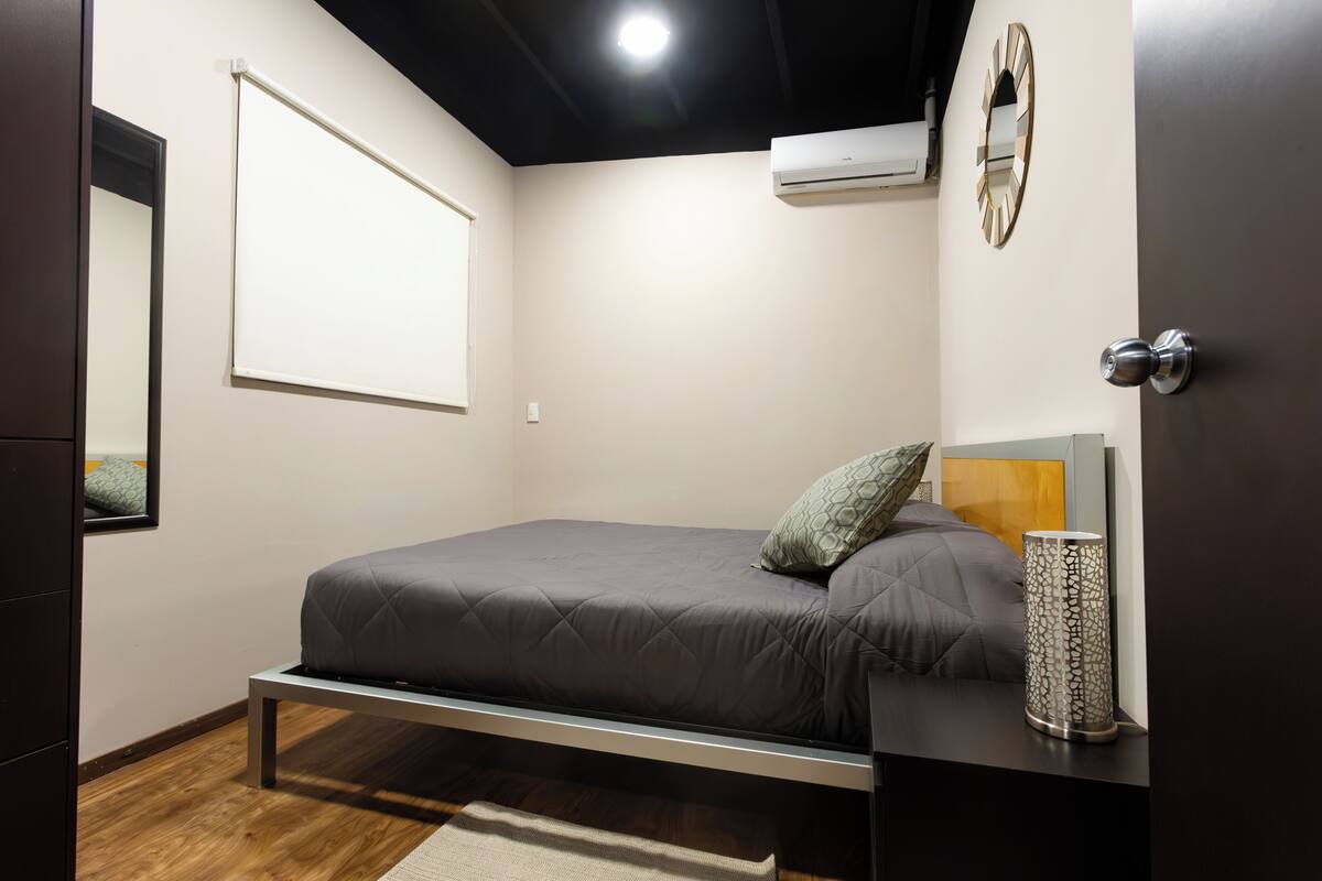 A modern bedroom with a bed, a bedside table, a wall-mounted air conditioner, a mirror, and a door.