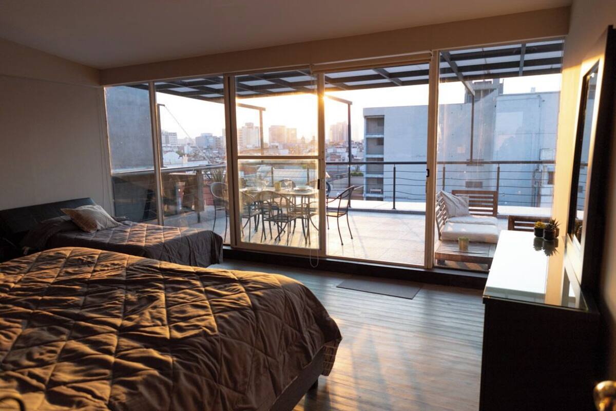 A bedroom with a large bed, a desk, and a balcony with a table and chairs.