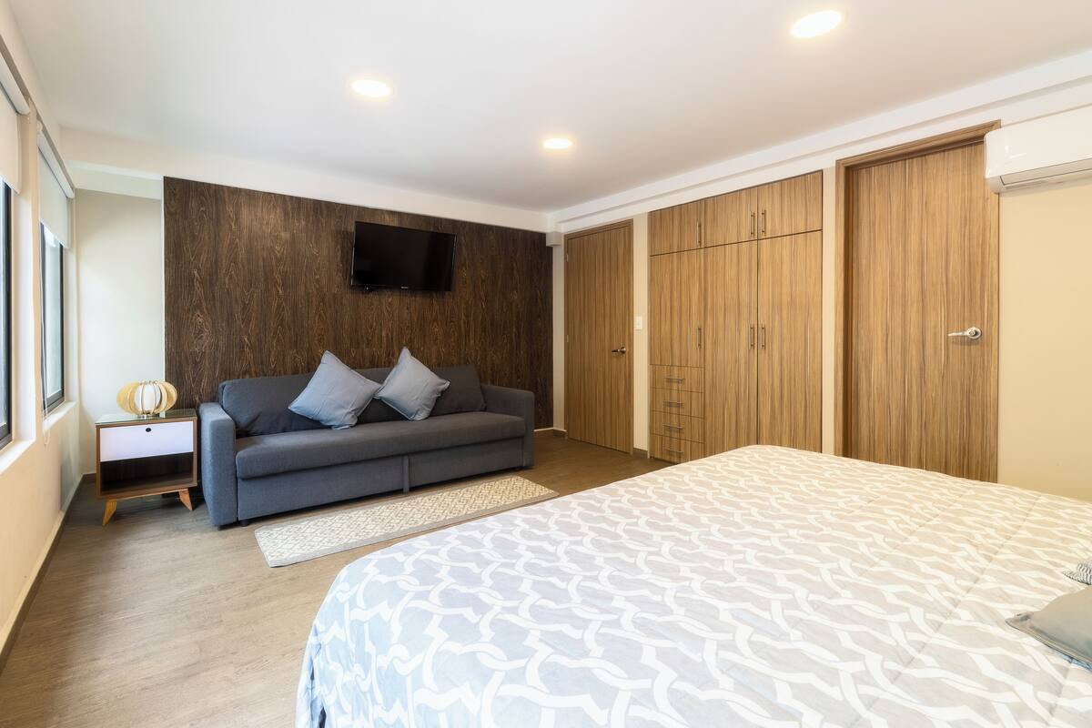 A bedroom with a bed, a grey sofa, a TV mounted on the wall, wooden wardrobes, and a small bedside table with a lamp.
