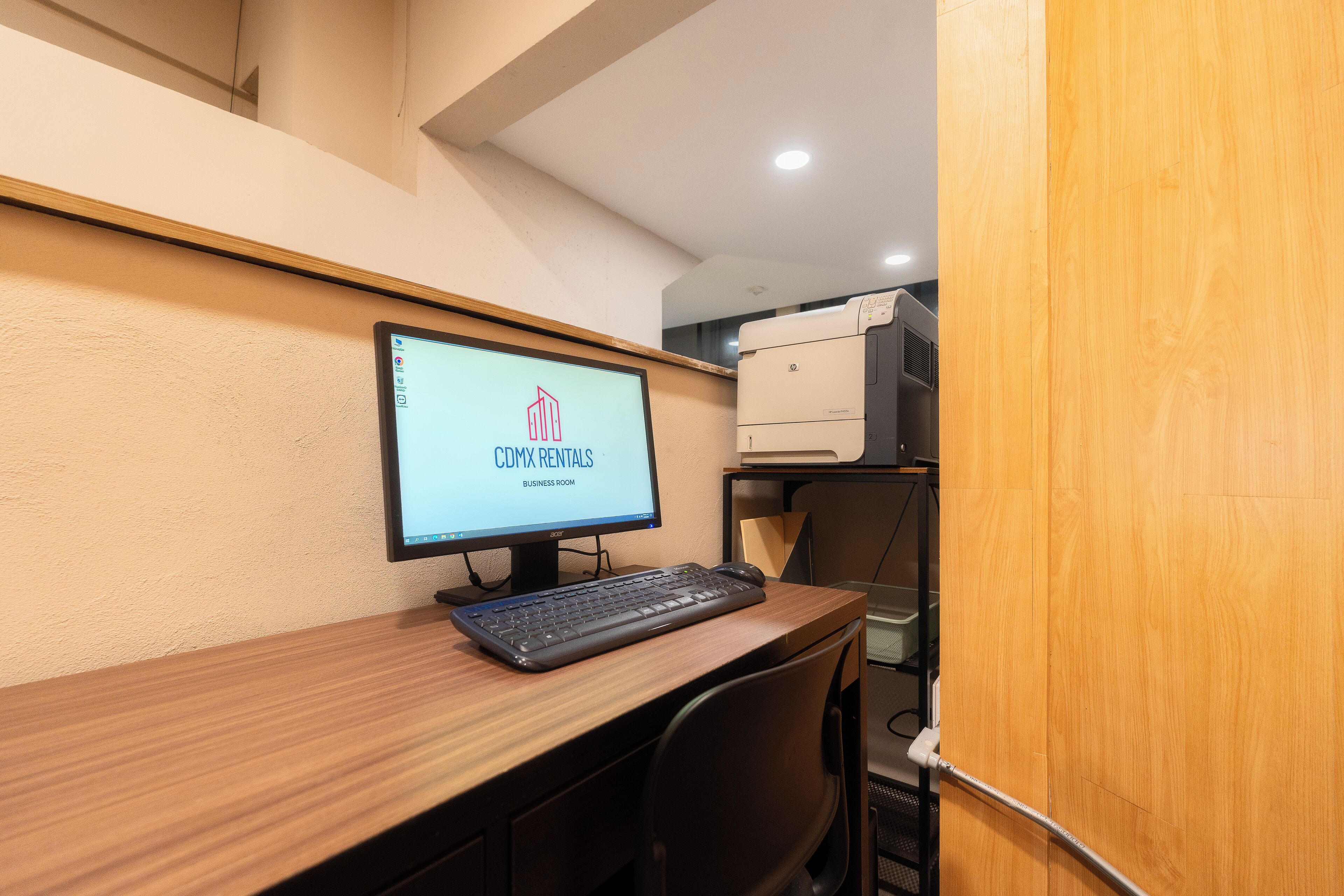Una habitación de negocios con un monitor de computadora mostrando 'CDMX Rentals', un teclado y una impresora sobre un escritorio de madera.