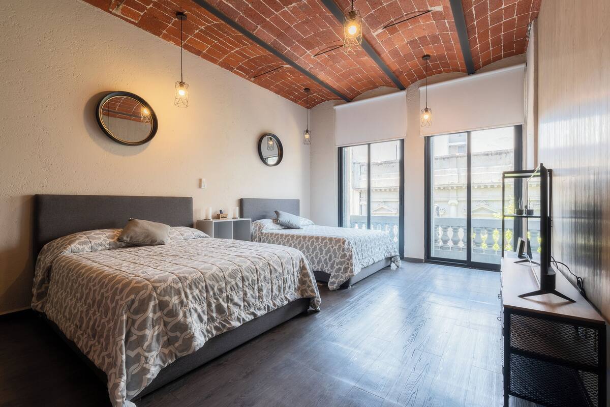 A modern hotel room with two beds, a large window, and a brick ceiling.