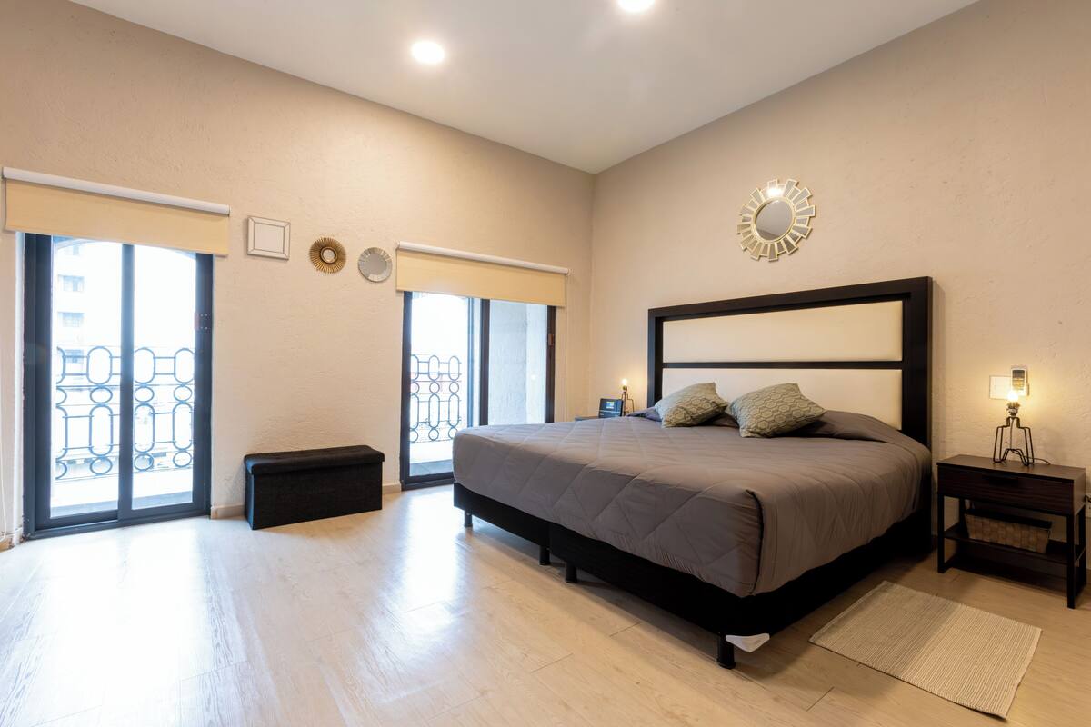 A bedroom with a large bed, a nightstand, a bench, and a window with a view of a balcony.