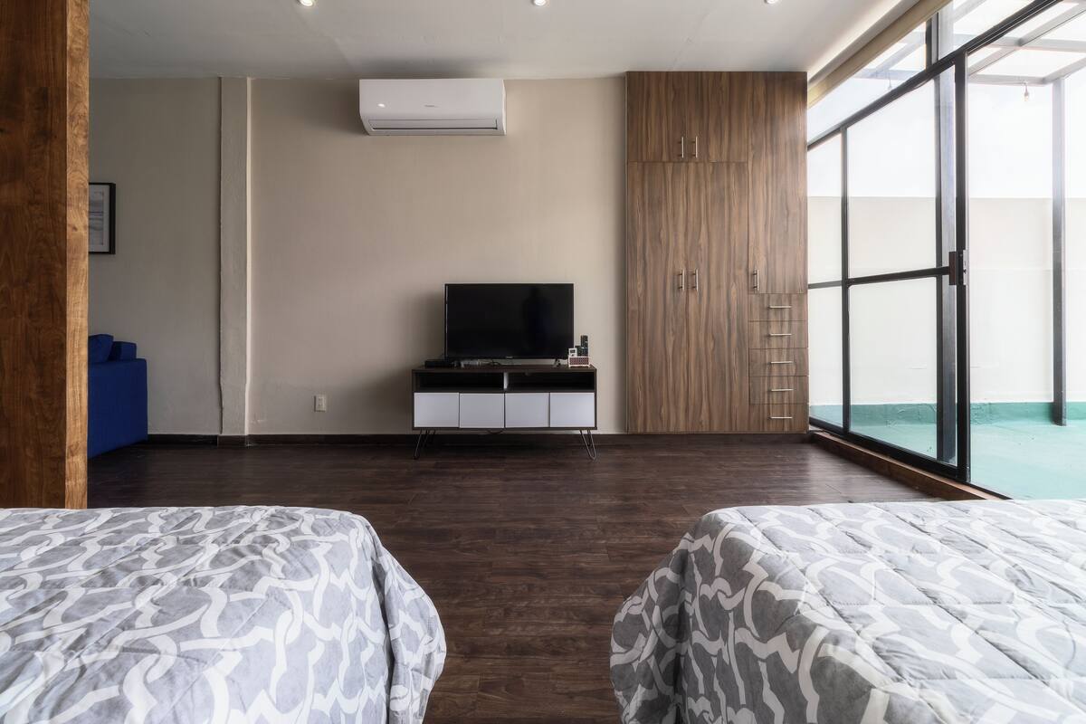A modern hotel room with a large window, a flat-screen TV on a white cabinet, and two beds with patterned bedding.