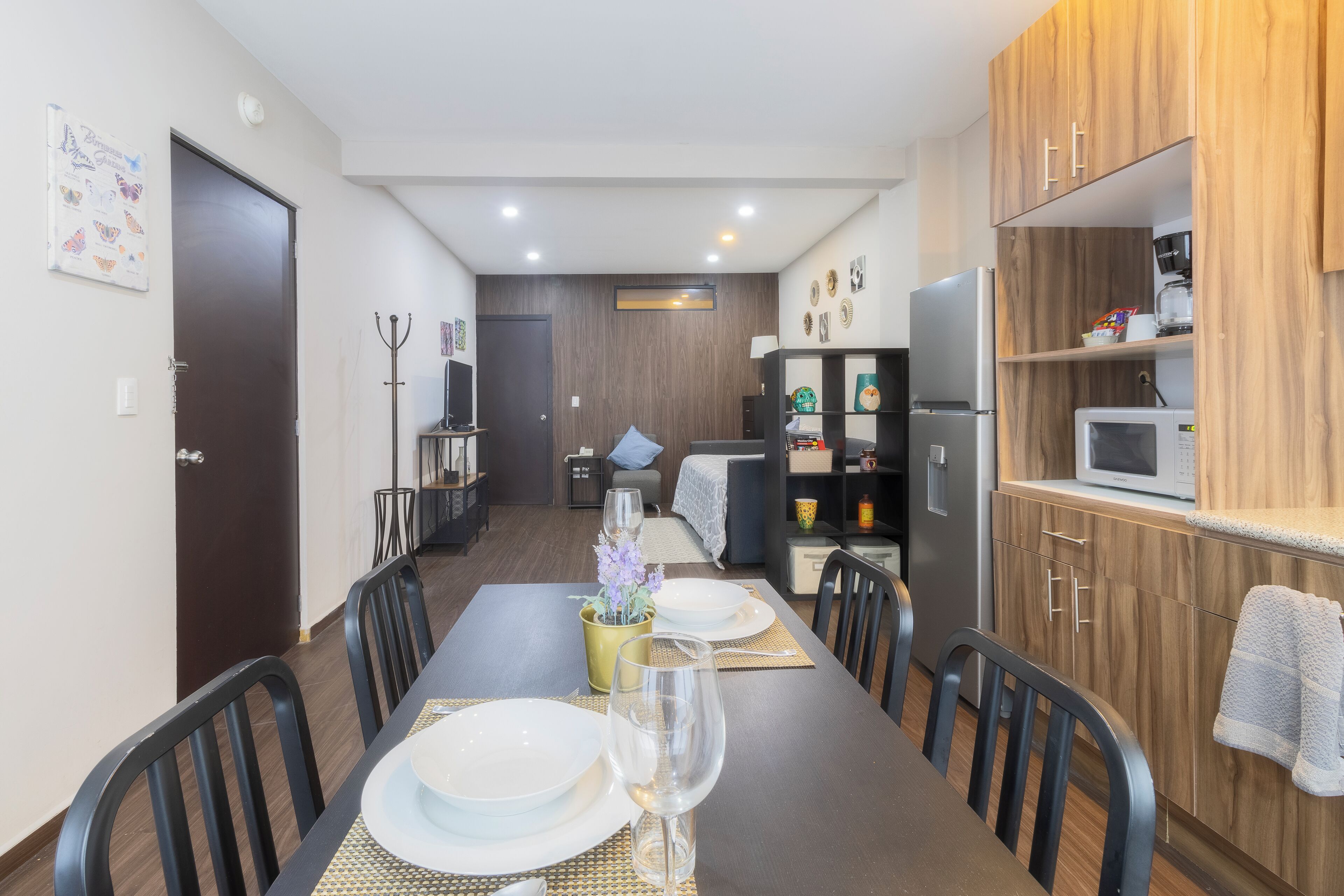 A modern kitchen with wooden cabinets, a microwave, and a coffee maker. There is a dining area with a table set for two, chairs, and a vase with flowers.