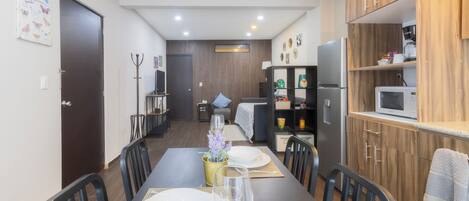 A modern kitchen with wooden cabinets, a microwave, and a coffee maker. There is a dining area with a table set for two, chairs, and a vase with flowers.