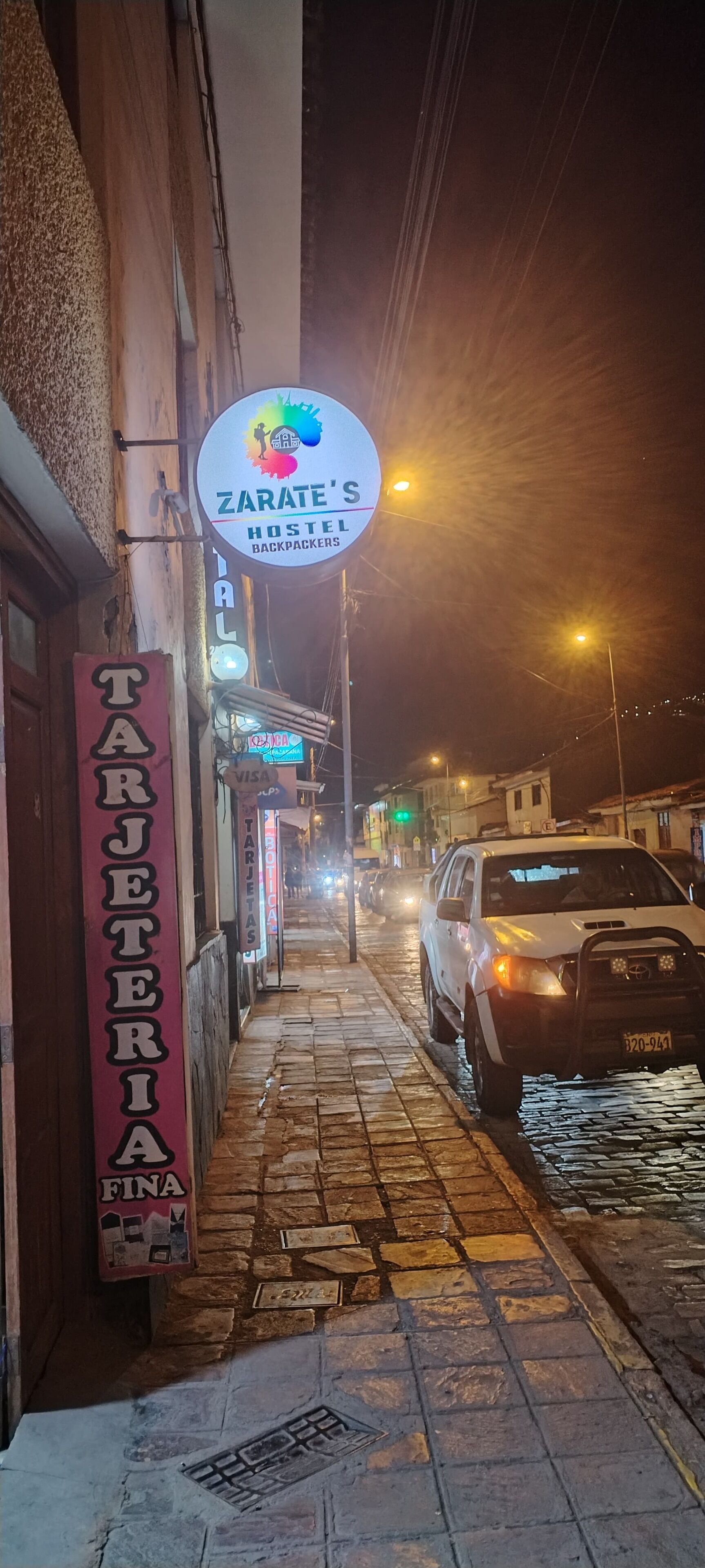 A narrow street with a hostel sign, a sign for a bar, and a parked car.