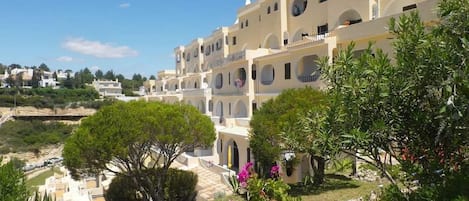 A multi-story building with balconies, surrounded by greenery and a clear sky.