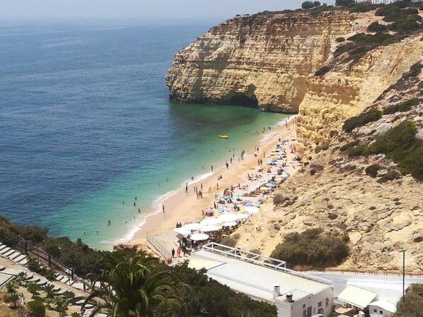 Ein Küstenstrand mit einer felsigen Klippe, einem Sandstrand und einer Reihe von Sonnenschirmen.