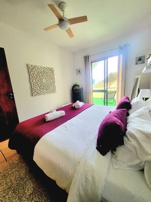 Ein Hotelzimmer mit Bett, Deckenventilator, Wanddeko, Fenster mit Vorhängen und Blick ins Freie.