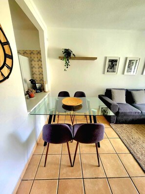 Ein Essbereich mit Glastisch und Stühlen, einer Couch, einem Teppich und einem an der Wand befestigten Regal mit einer Pflanze.