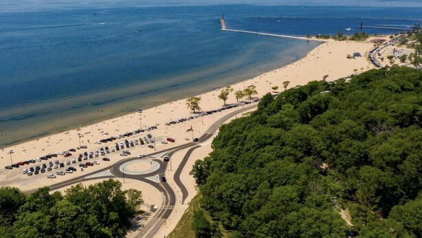 Vista aérea de una zona costera con playa, un faro y una carretera serpenteando por el paisaje.