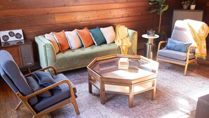 Una habitación acogedora con un sofá verde, dos sillones, una mesa de centro de vidrio, suelo de madera y una pared de madera.